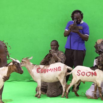 Still from Once Upon a Time in Uganda. Four men in front of a greenscreen outside. One is standing, holding a camera. Two men kneel on the ground holding guns and two goats, the words "Cooming soon" are painted in red on the two goats. Another man crouches above a goat with a red logo painted on it.