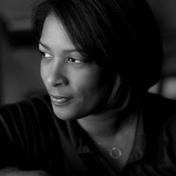 Dawn Porter has short hair, wears a necklace, and a long sleeve v-neck blouse. She smiles with her face looking towards the side. Black and white portrait.
