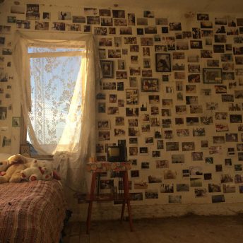 A still from The Guardian of Memory. The interior of a bedroom. One wall has a window and the rest of the wall is covered in photos.