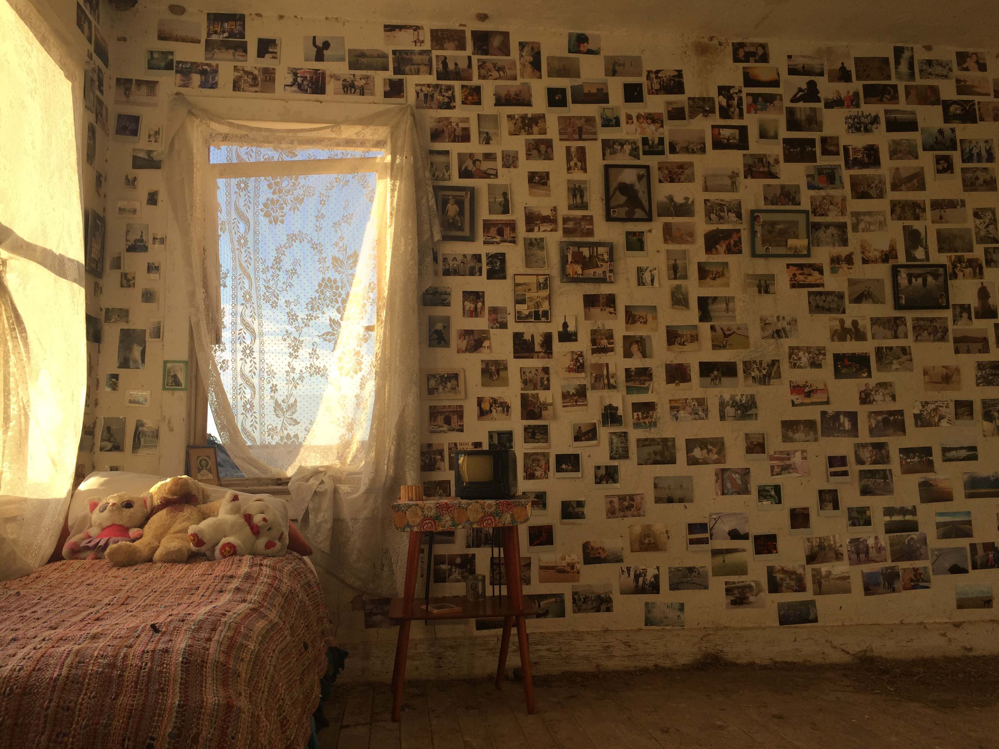 A still from The Guardian of Memory. The interior of a bedroom. One wall has a window and the rest of the wall is covered in photos.