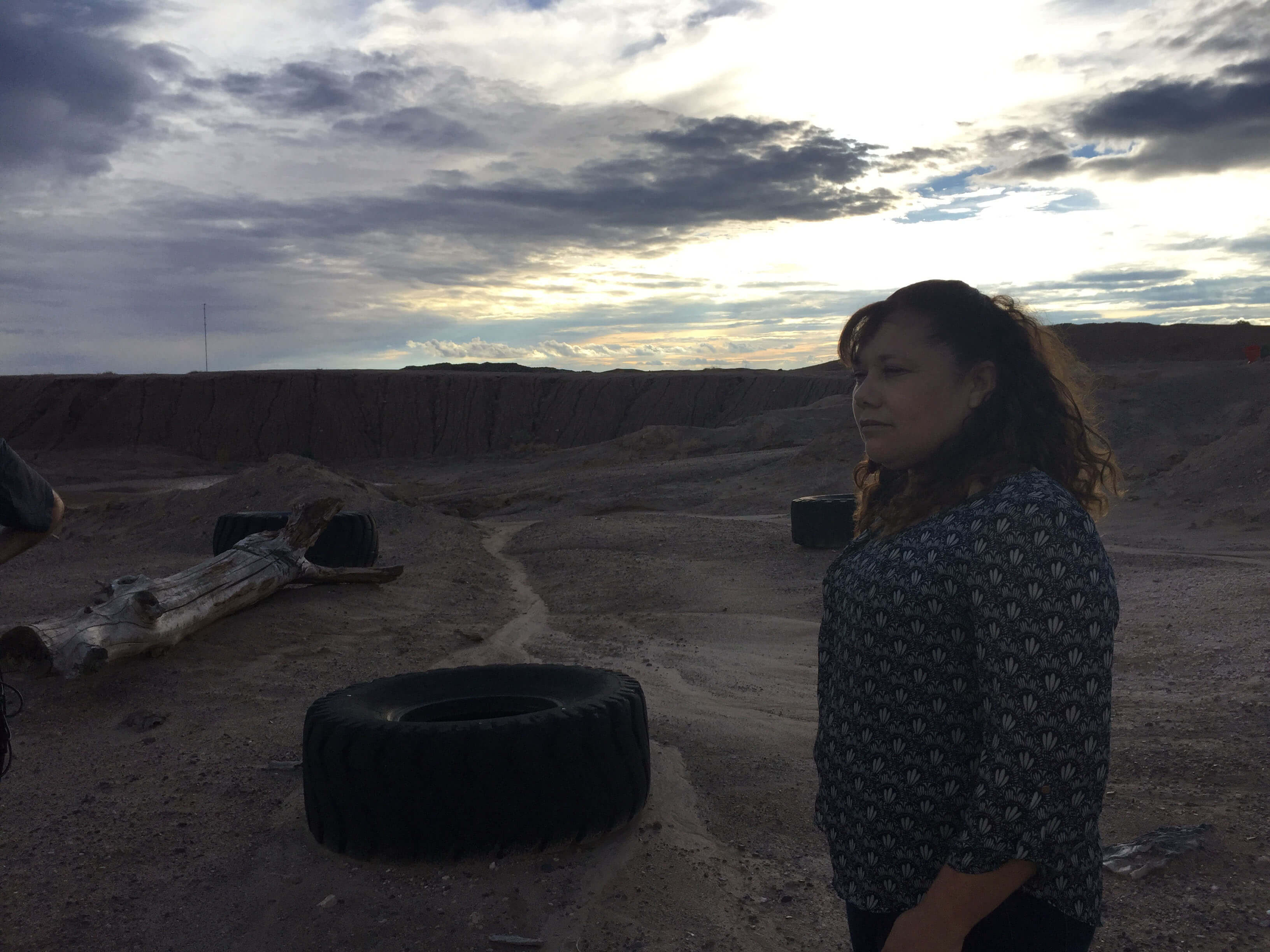 A still from The Guardian of Memory. A photo of a person in a dirt valley, looking off into the distance. There is a tire on the ground nearby.