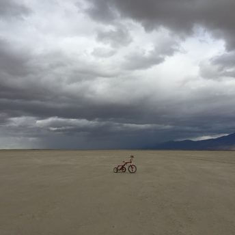 A still from The Guardian of Memory. A red tricycle sits in the middle of a desert with large mountains off in the distance. The sky is cloudy.