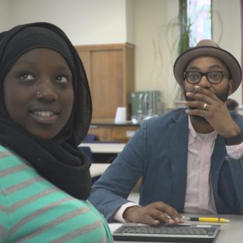 Still from Kids Can Spit. A female high school student in a hijab is looking out the frame, while behind her, a male teacher looks in the same direction. He is typing on a keyboard with one hand, and his other hand covers his mouth. They are in a science classroom.