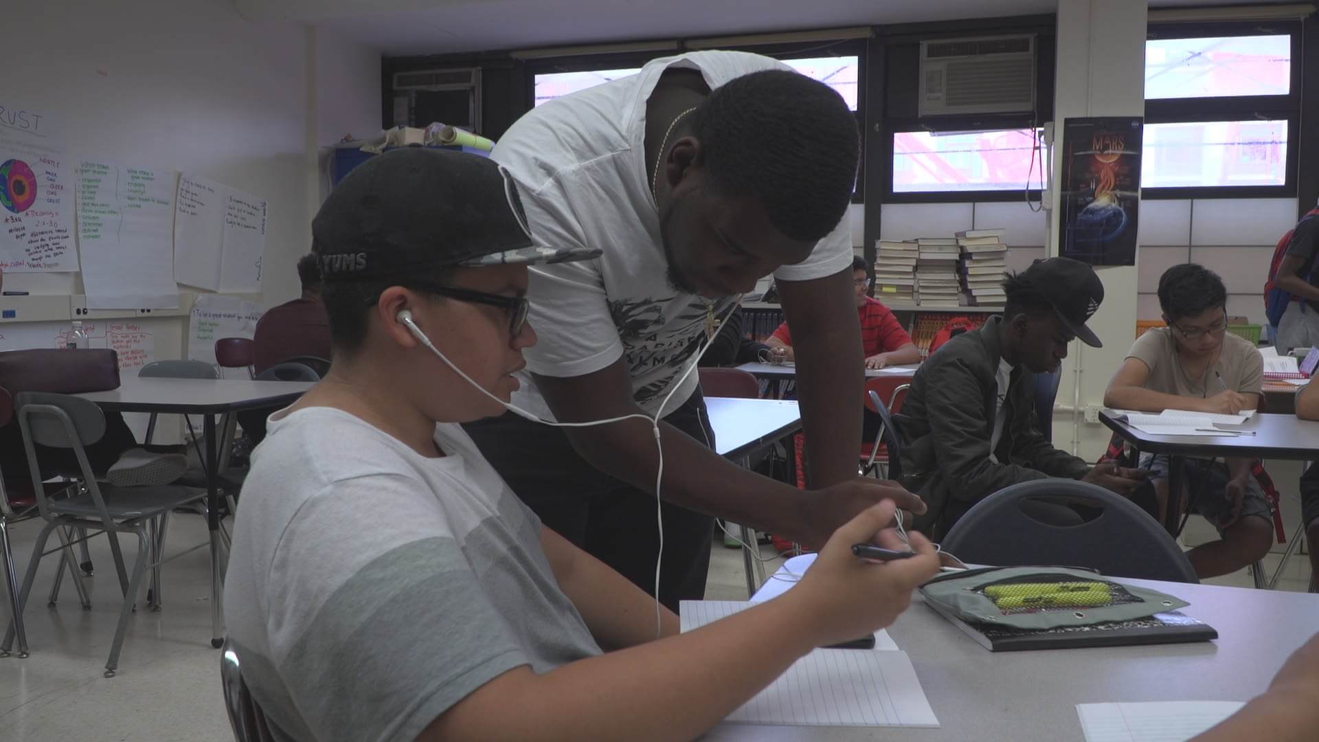 Still from Kids Can Spit. Two male high school students share wired earbuds. One student is sitting at a desk and holds a pen as he looks at a lined white sheet of paper. The other student is standing upright and leaning over the desk, also looking at the paper. Behind them, other students are sitting at desks.