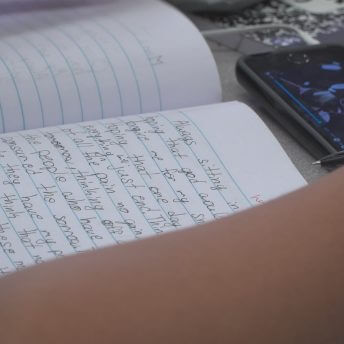 Still from Kids Can Spit. Close up of a wide-ruled notebook with full page of handwritten text. An arm rests on a table to the side of the notebook, and a smartphone is on the table to the top of the notebook, playing a music video of a man wearing headphones and singing.