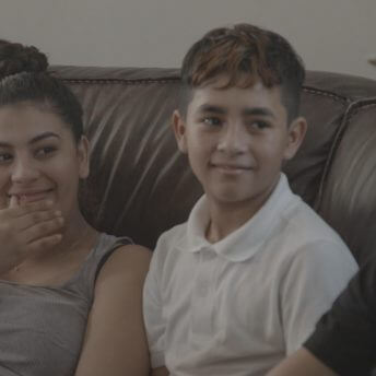 A smiling woman in a grey tank next to a young boy in a white collared shirt, sitting on a sofa and looking to their left.