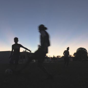 Sunset silhouette of three young people in motion playing soccer outdoors on grass.