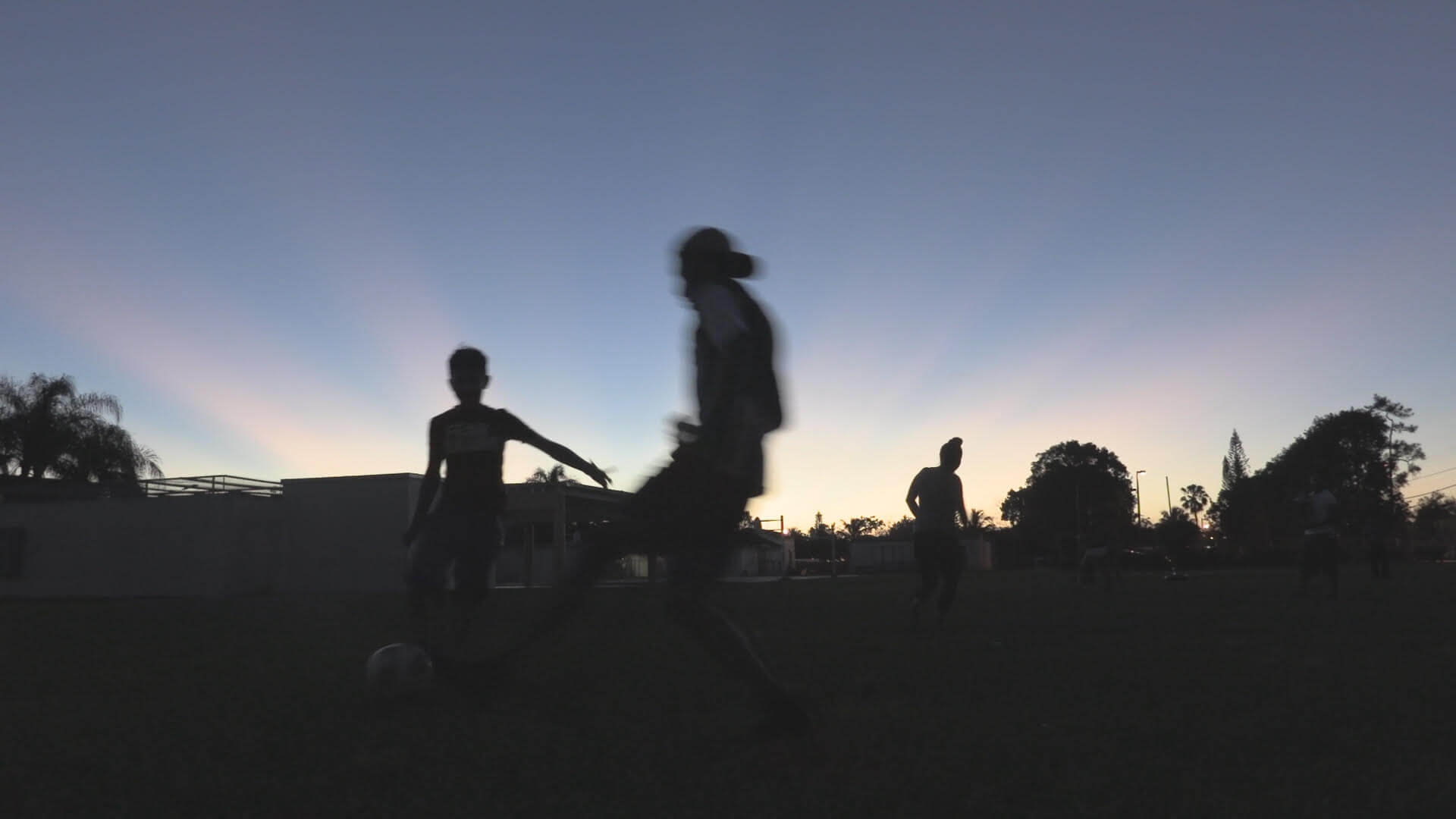 Sunset silhouette of three young people in motion playing soccer outdoors on grass.