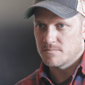 A man looks away from the camera. He wears a plaid shirt and a cap.