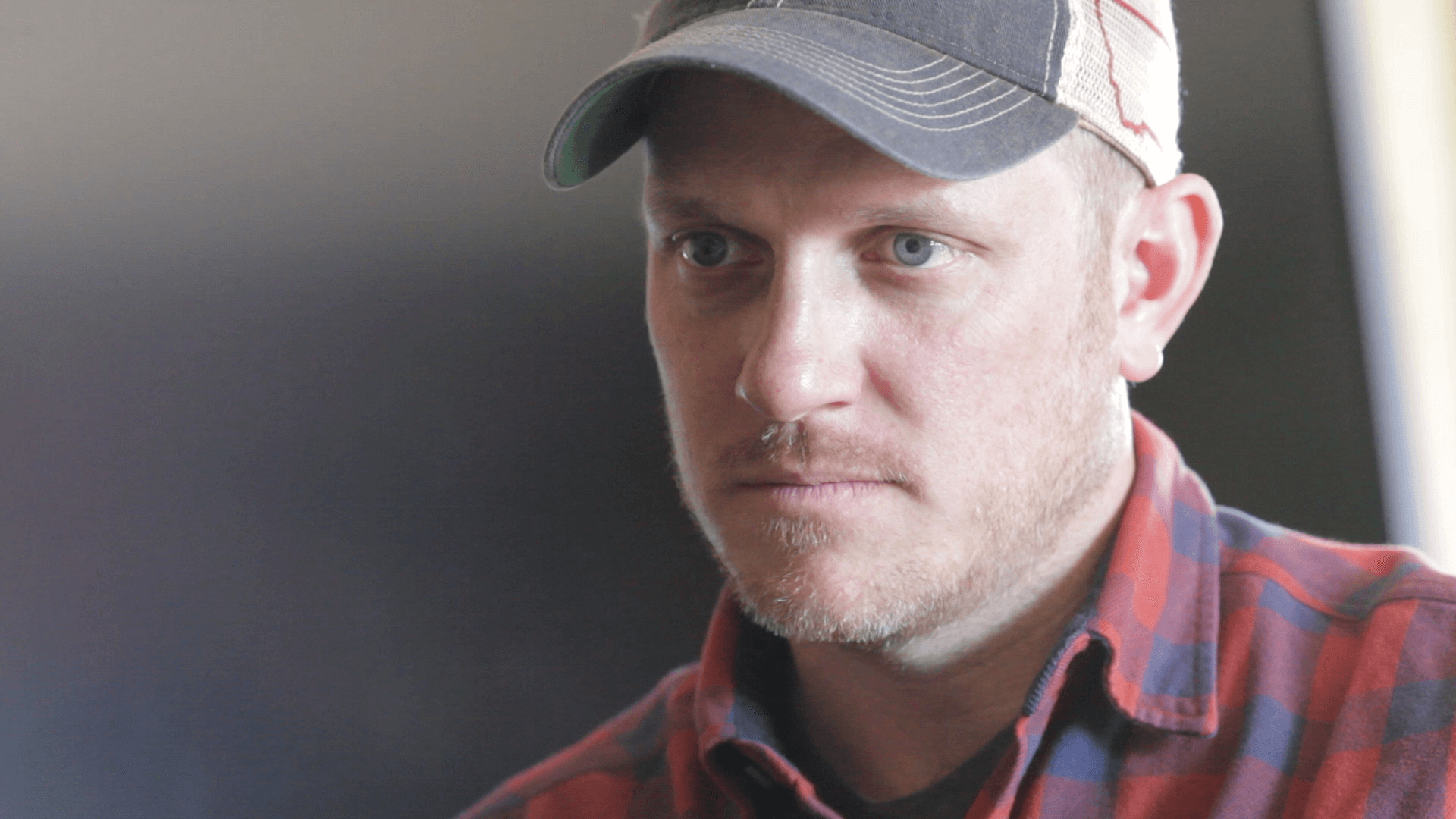 A man looks away from the camera. He wears a plaid shirt and a cap.
