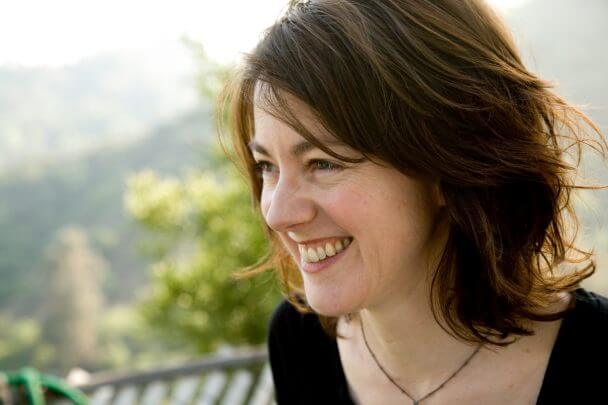 Laura Nix profile, smiling. She has shoulder-length wavy brown hair, wears a black top, and a necklace. Out of focus trees and sunny background.