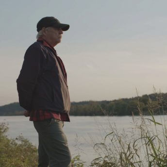 A still from The Devil We Know. A man wearing a black hat, jacket, and blue jeans stands on the edge of a body of water. He looks off into the distance. It is daytime.