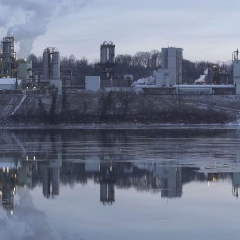 A still from The Devil We Know. A photo of a body of water. Across the water sits an industrial plant with smoke rising from columns.