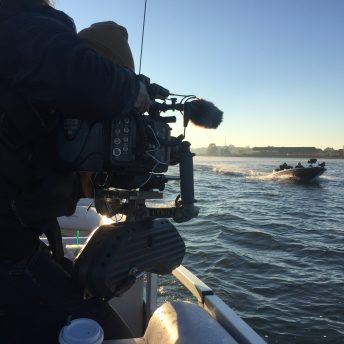 A production still from The Devil We Know. A person operating a large camera mounted to the side of a boat. They are pointing the camera at another boat passing by on the water, headed in the opposite direction. It is daytime.