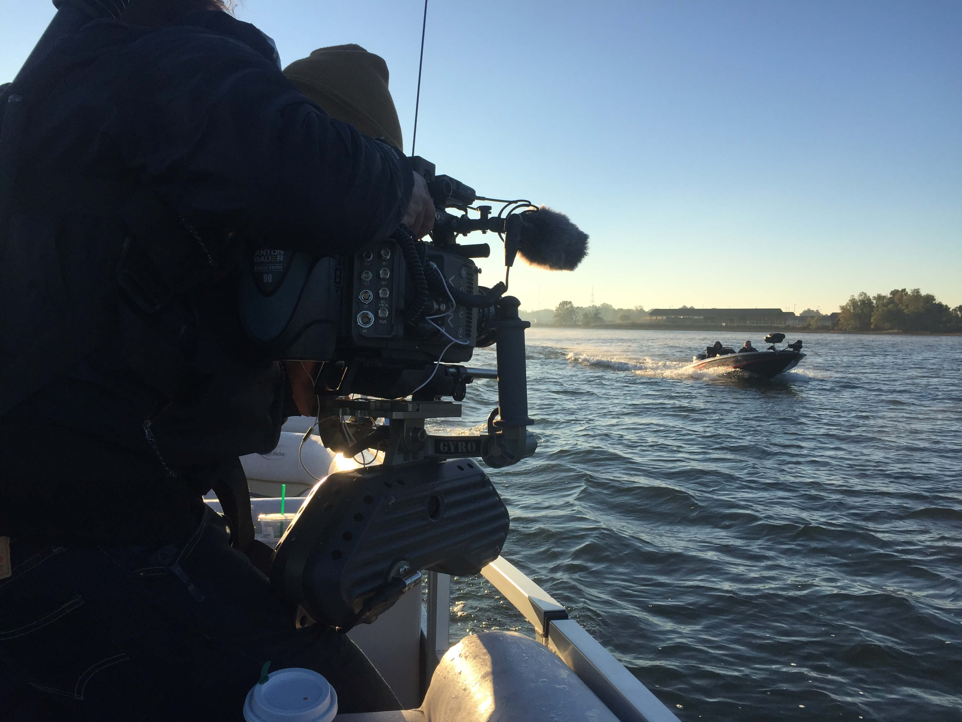 A production still from The Devil We Know. A person operating a large camera mounted to the side of a boat. They are pointing the camera at another boat passing by on the water, headed in the opposite direction. It is daytime.