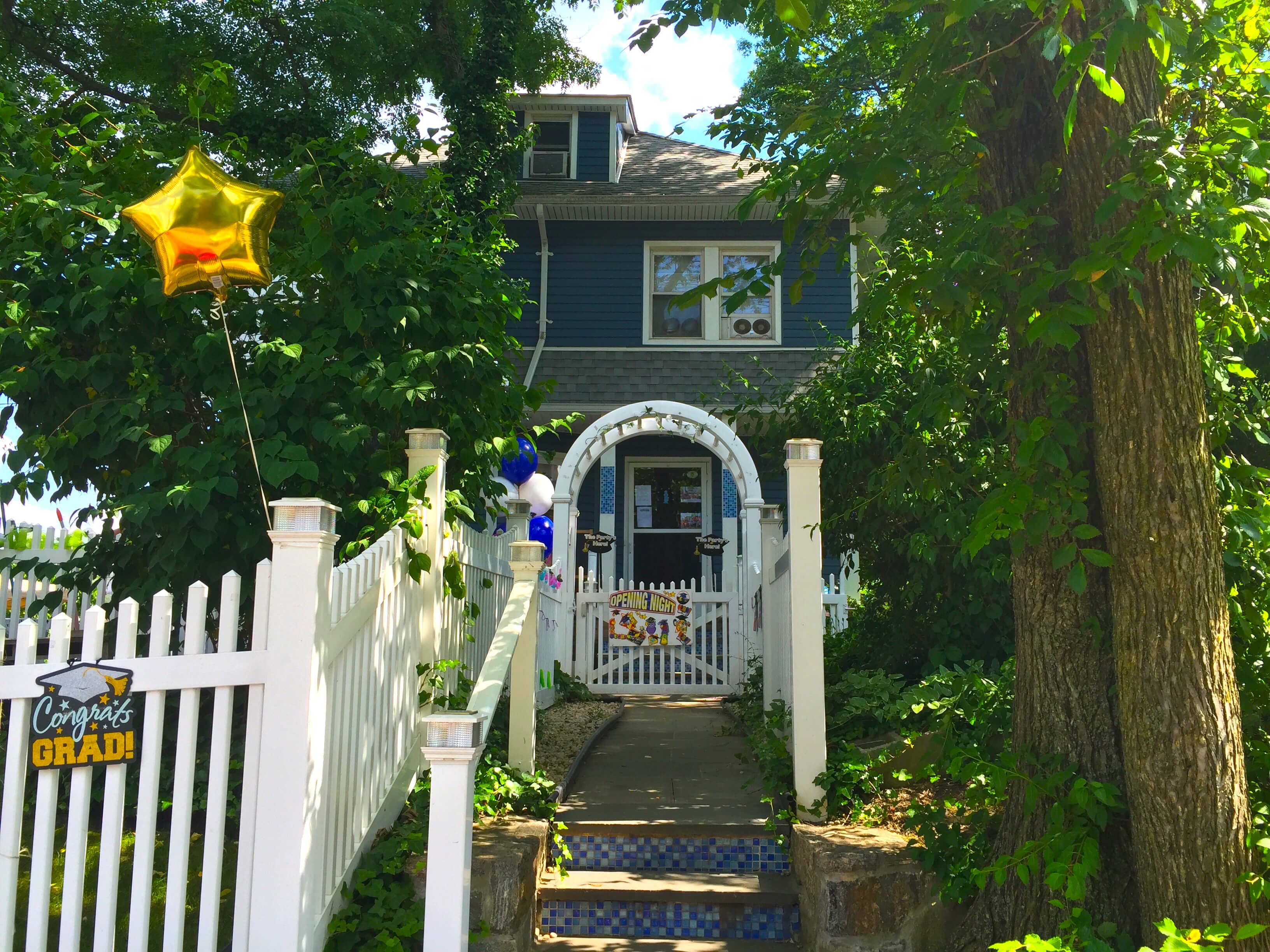 An Exterior shot of a blue house surrounded by green vegetation. There is a balloon in the shape of a golden star tied to the white fence in front of the house.
