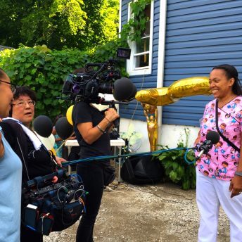 A camerawoman and a soundperson are interviewing a woman in a pink uniform. They are all standing in front of a blue house.