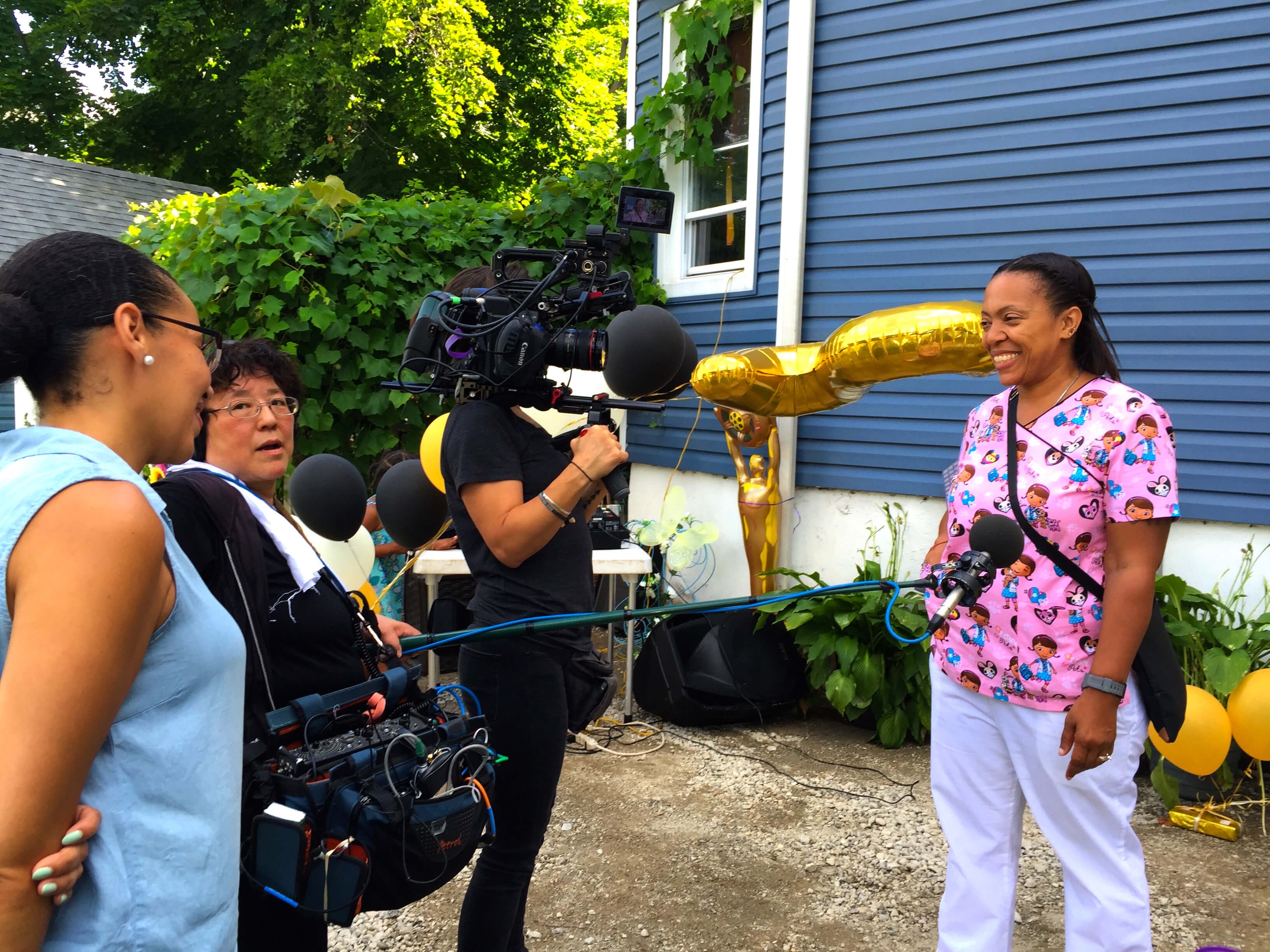 A camerawoman and a soundperson are interviewing a woman in a pink uniform. They are all standing in front of a blue house.