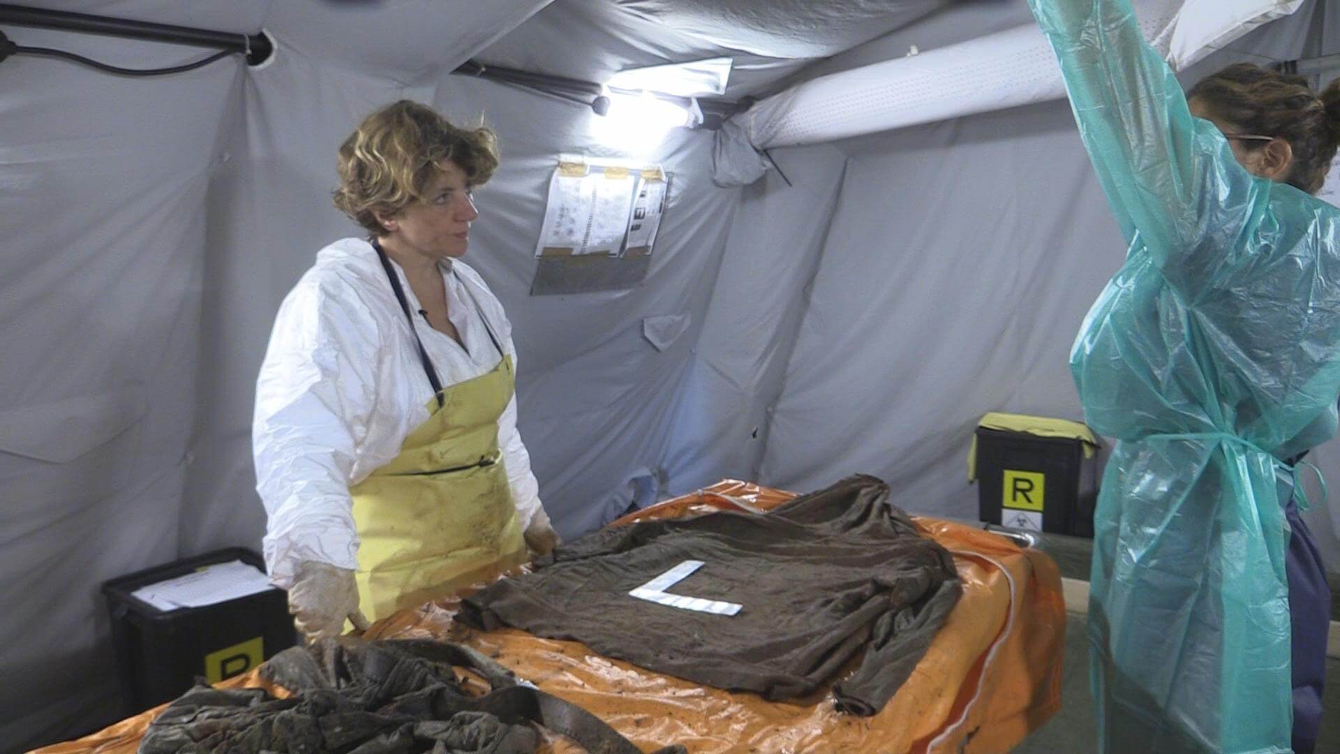 Still from Number 387. Two women in a medical tent stand at a table across from each other wearing PPE. One woman has a hand raised. There is an item of clothing on the table with an "L" ruler on top of it.