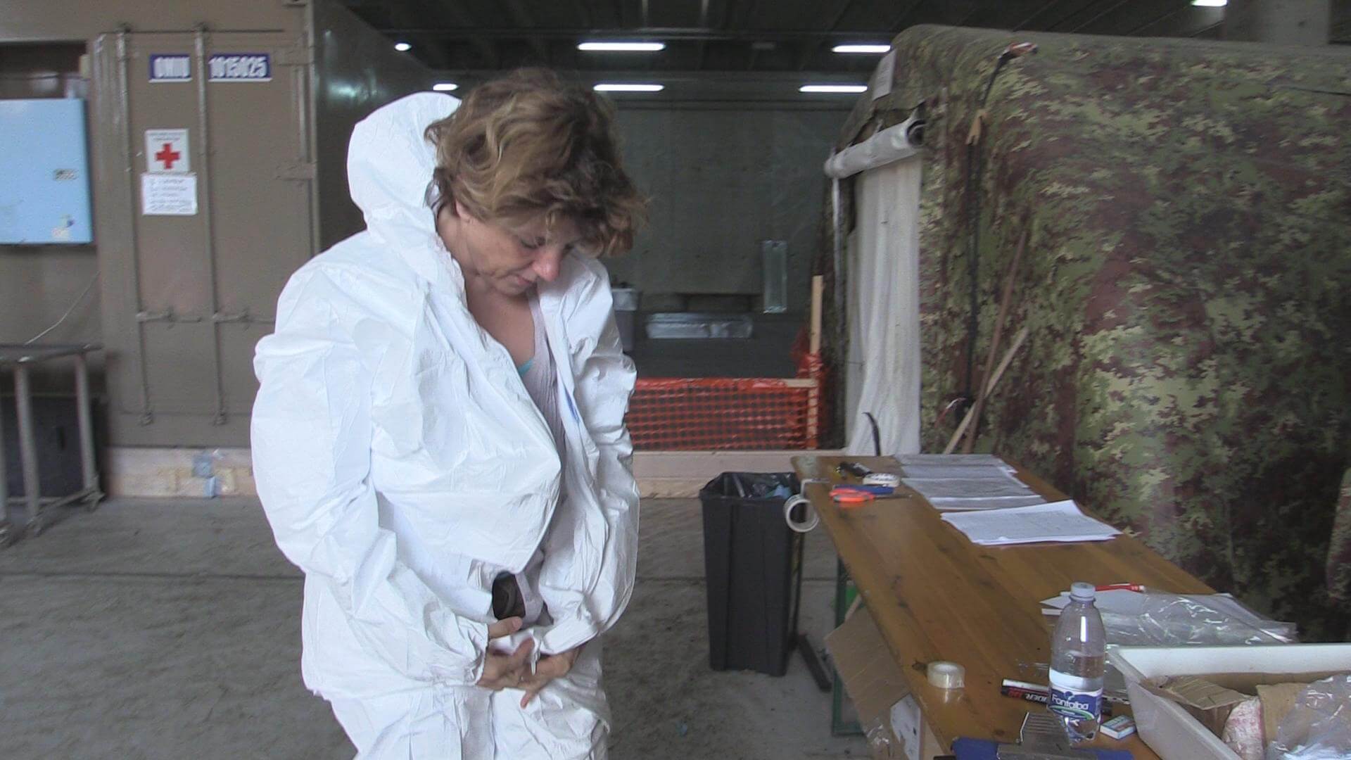 Still from Number 387. A woman zips up her medical hazard suit. She is standing in front of a table and military tent structure.