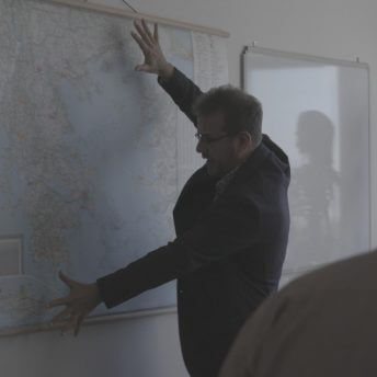 A man in a suit stands in front of a big map hanging on the wall. He spreads his hands.
