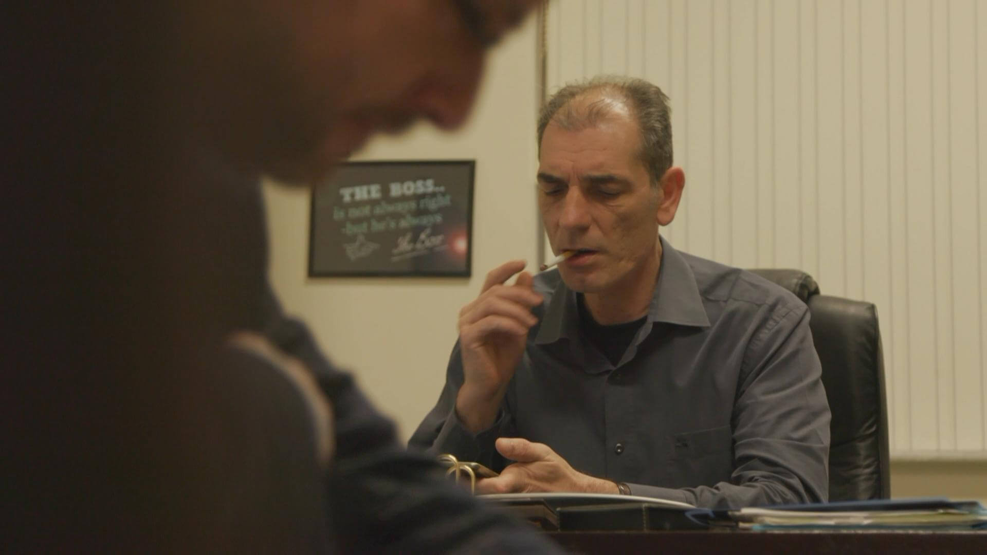 Still from Number 387. A man is sitting at a desk smoking a cigarette and holding a cell phone. There is an out of focus person in the foreground.