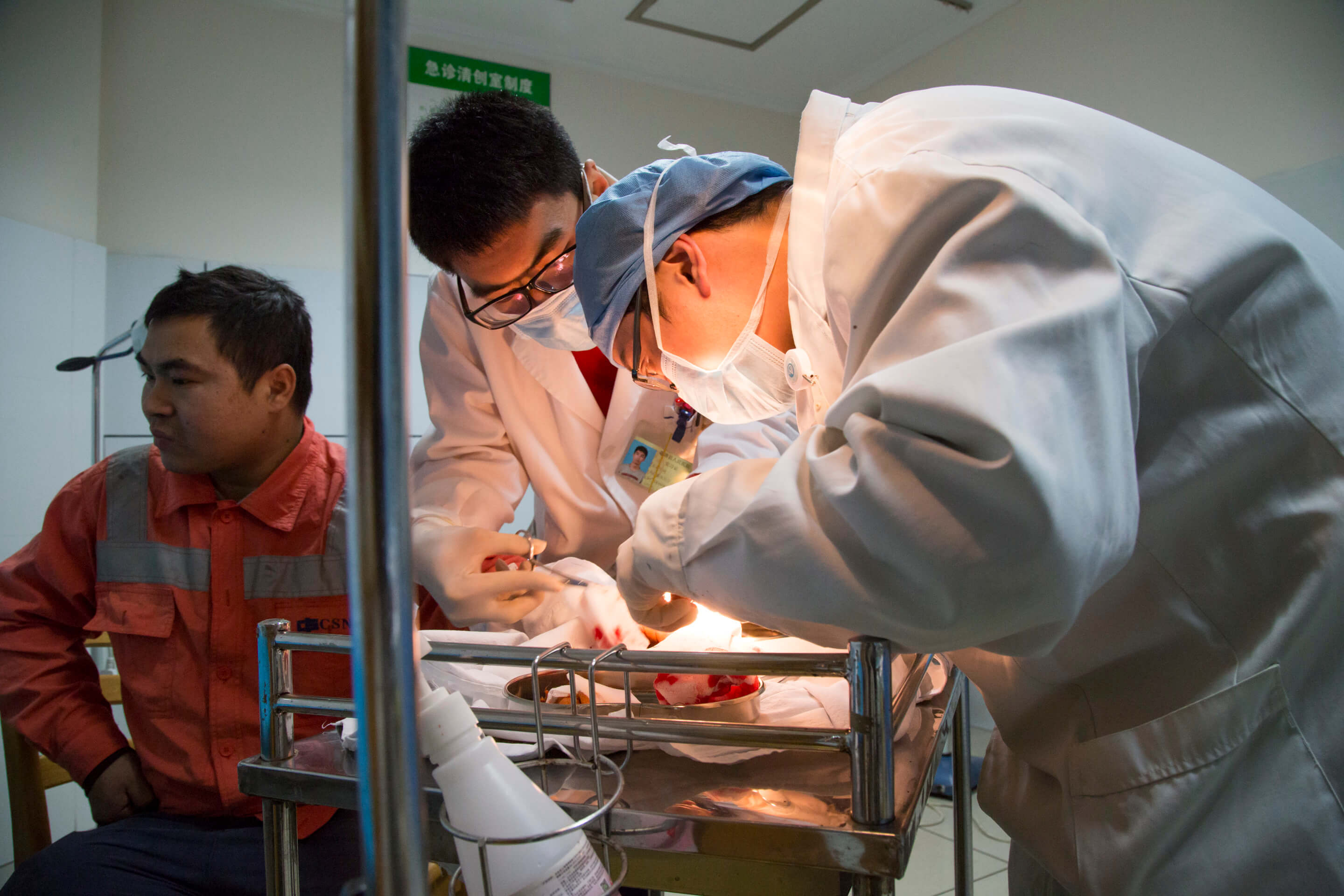Two medical staff in lab coats hover over a brightly lit tray of bloody gauze and a man in an orange jacket sits nearby