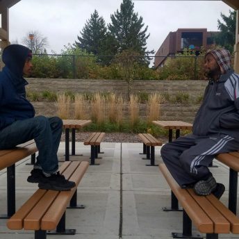 Profile full shot of two men sitting in front of each other on park benches. Both of them wear jackets and have their hands inside their pockets.