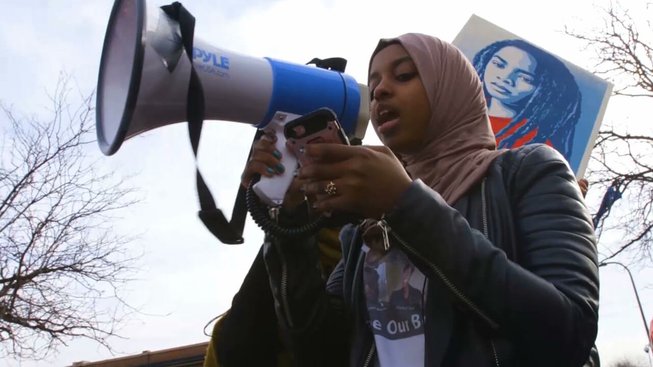 Still from Accept the Call. Low angle shot of a young girl speaking through a megaphone while reading from a cellphone. She is wearing a hijab, a t-shirt, and a jacket.