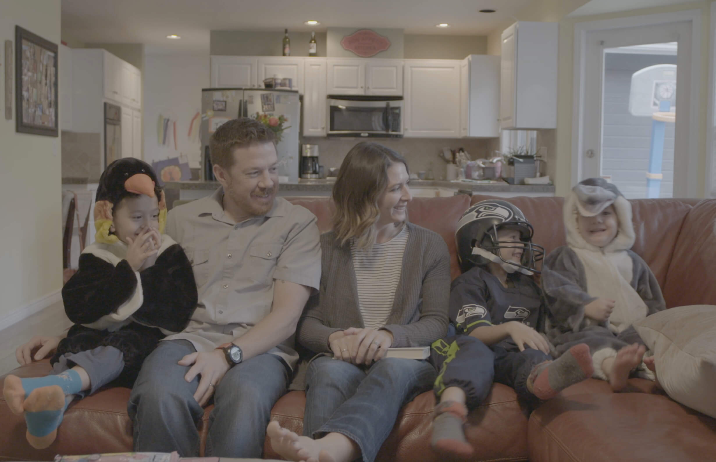 Still from Made in Boise. A man, woman, and three children in costumes sit on a couch, smiling. There is an out of focus kitchen in the background.
