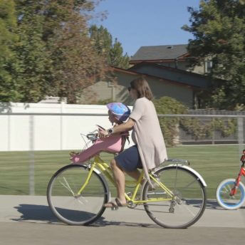 Still from Made in Boise. A woman is outside riding a bike down the sidewalk of a neighborhood street.