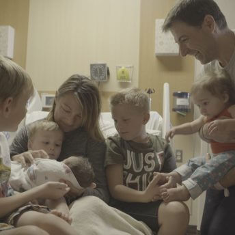 Still from Made in Boise. A woman sitting up in a hospital bed is holding an infant and toddler on her lap. There are three other children sitting around her on the bed and a man standing next to the bed, holding a small child.