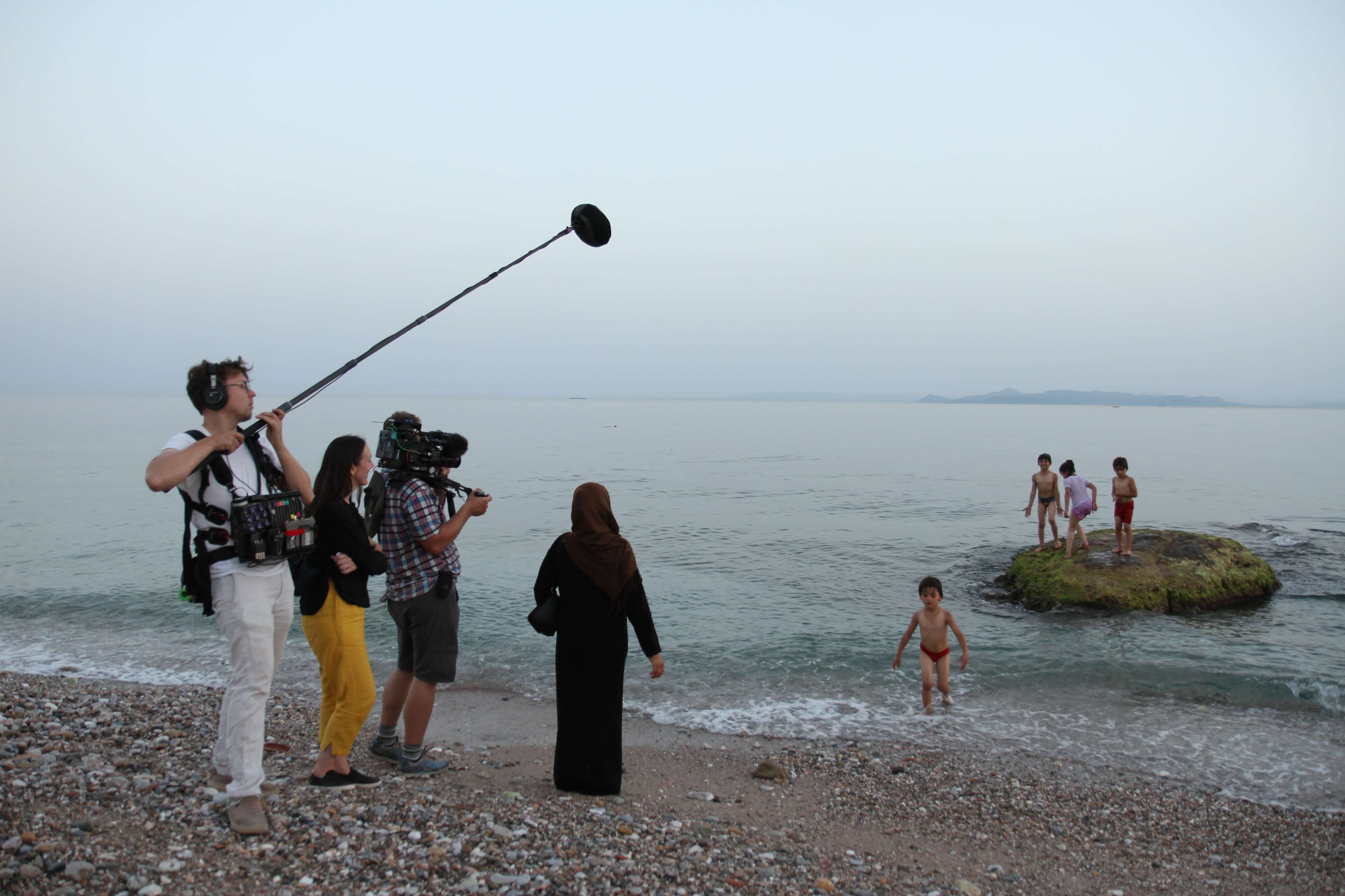 Production still from As Simple As Water. Director Megan Mylan, and her cameraman and audio person, follow a family playing on the ocean shore. The mother waits while the children play in the water.