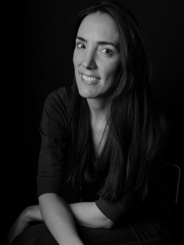 Megan Mylan looks directly at the camera reclining her hands on her knees and smiles widely. Black and white portrait.