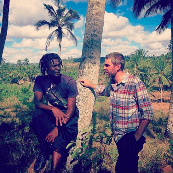 A production still from The Letter. Chris King stands with one of the documentary participants in the middle of a field populated with palm trees. It is sunny and the sky is blue.
