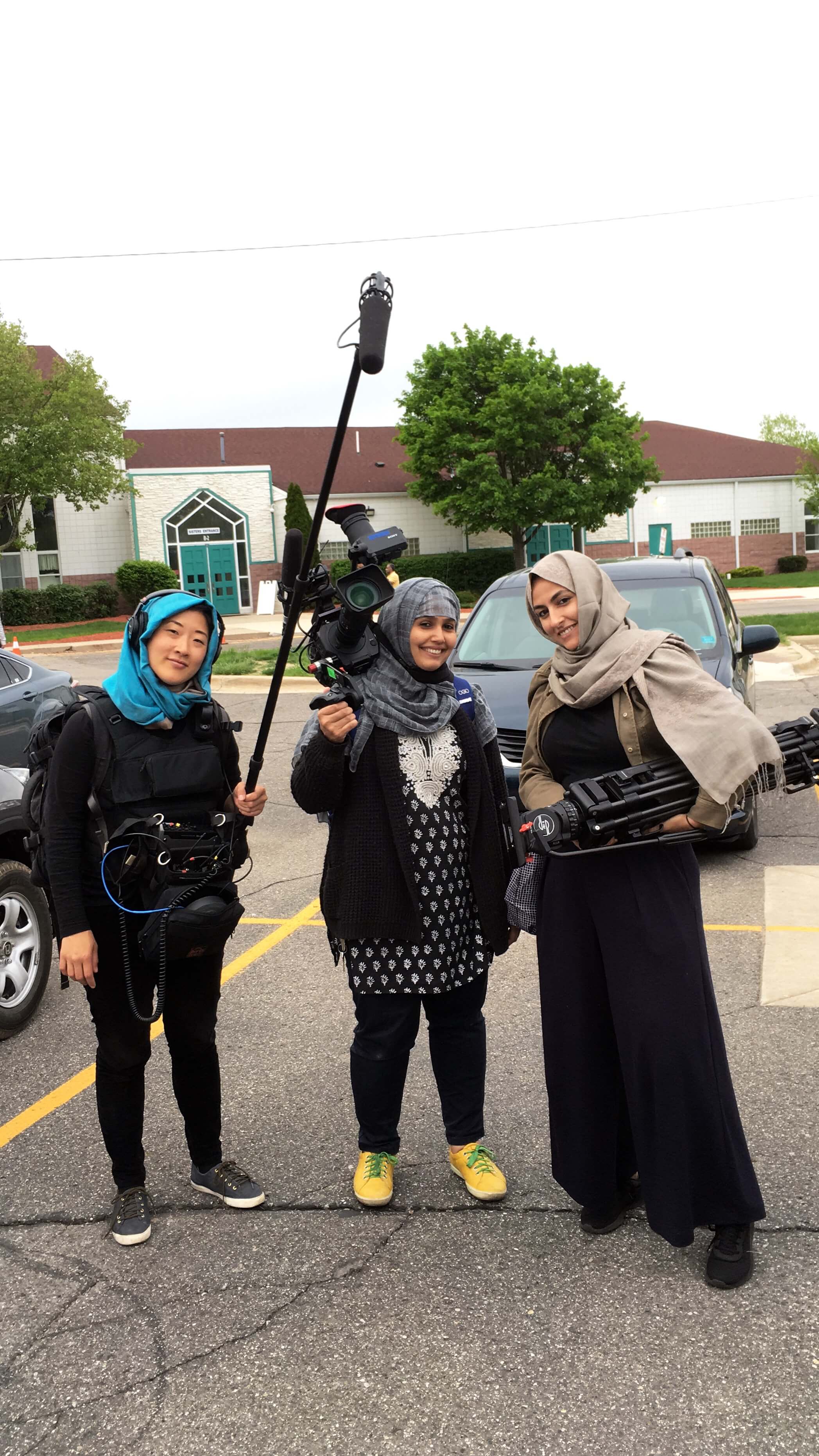 Production still of An Act of Worship. Three women crew members are standing in a parking lot, wearing hijabs, and holding film equipment.
