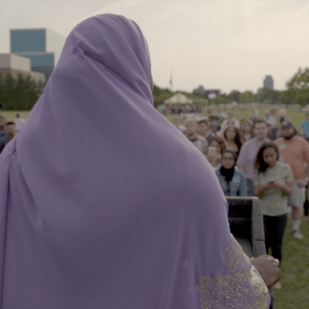 Still from An Act of Worship. Khadega Mohammed is standing at a podium and talking to a group of people. She is wearing a lilac Khimar.