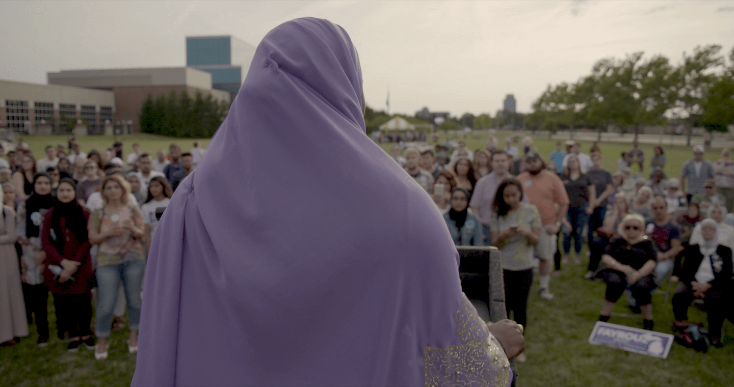 Still from An Act of Worship. Khadega Mohammed is standing at a podium and talking to a group of people. She is wearing a lilac Khimar.