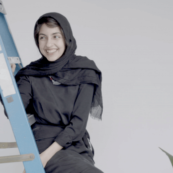 Still from An Act of Worship. A woman is sitting in a foldable chair, she is wearing a black dress, and a black hijad.