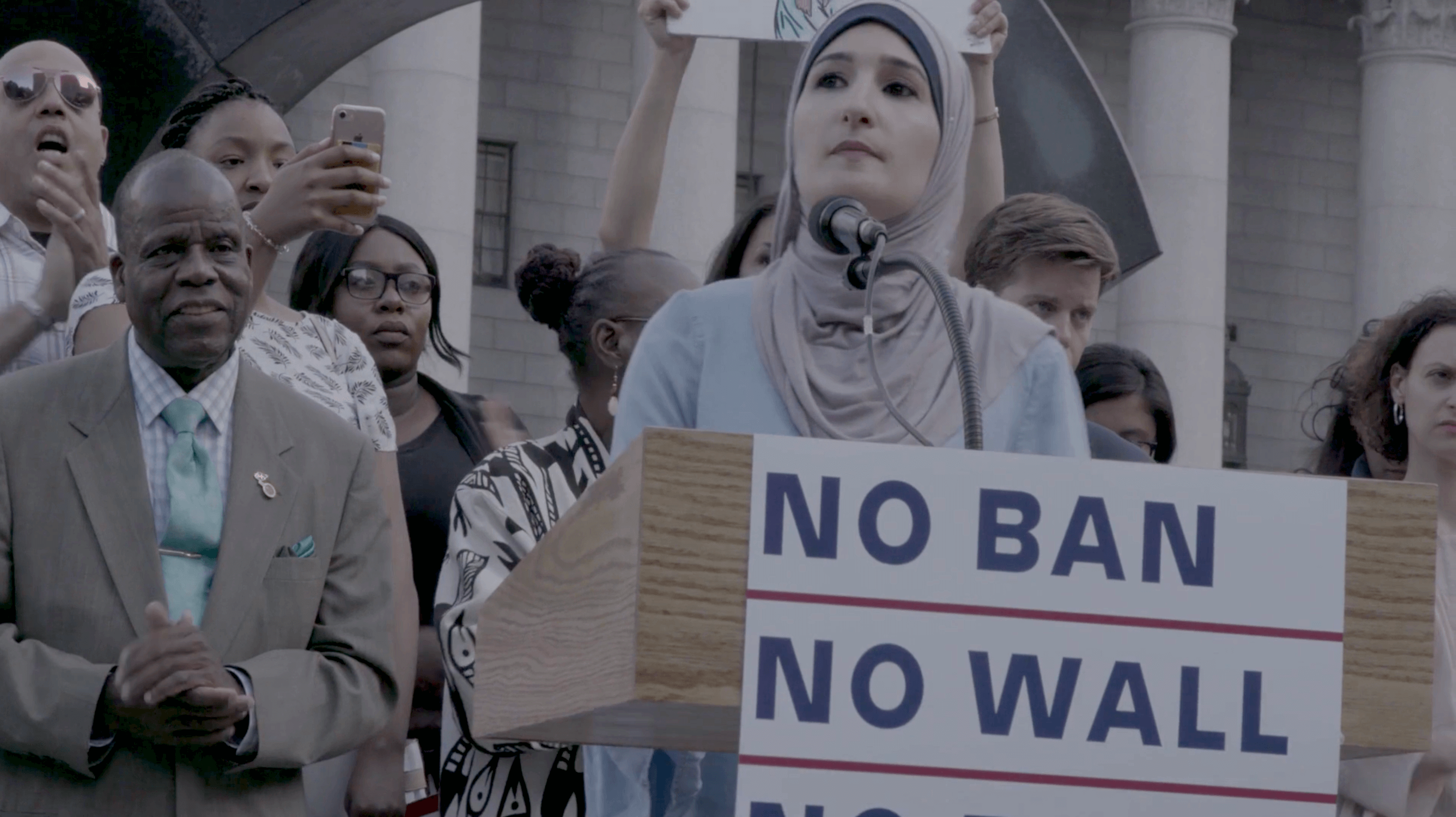 Still from An Act of Worship. A woman wearing a hijab is speaking in front of a podium, the podium has a poster that says "no ban, no wall." Behind her, there is a group of people.