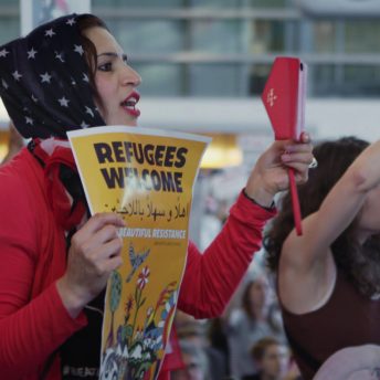 Still from An Act of Worship. People participating in a protest. A woman holds a poster that says "Refugees welcome".