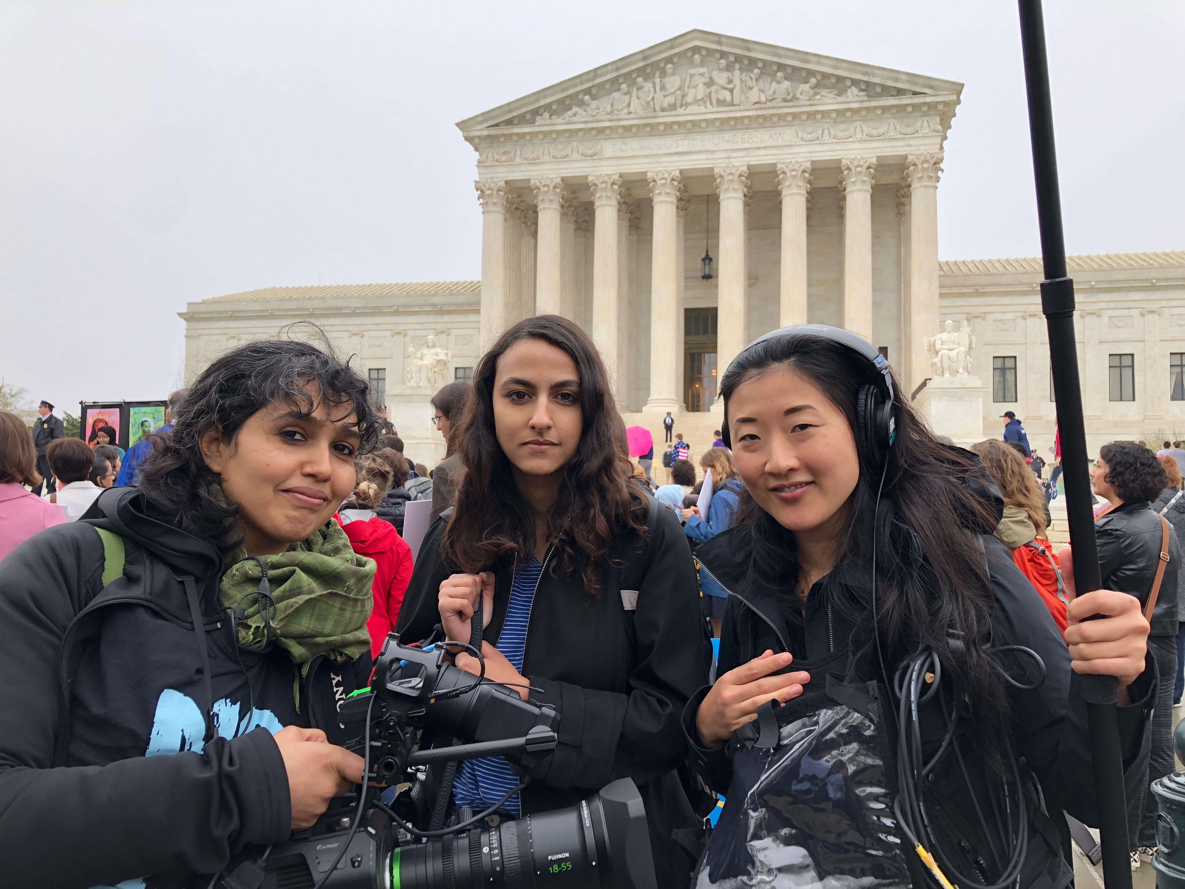 Production still of An Act of Worship. Three women dressed in black of the film's production crew are standing in front of the United States Supreme Court, director Nausheen Dadabhoy, to the left, is holding a camera.
