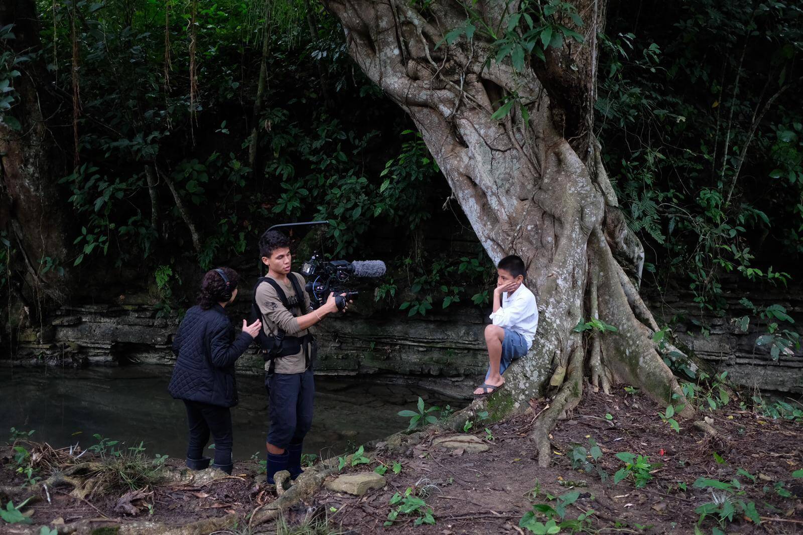 Production still from Silent Beauty. Two persons, one with a camera and the other with a headshot work recording a young boy leaning on a big tree. They are surrounded by nature.