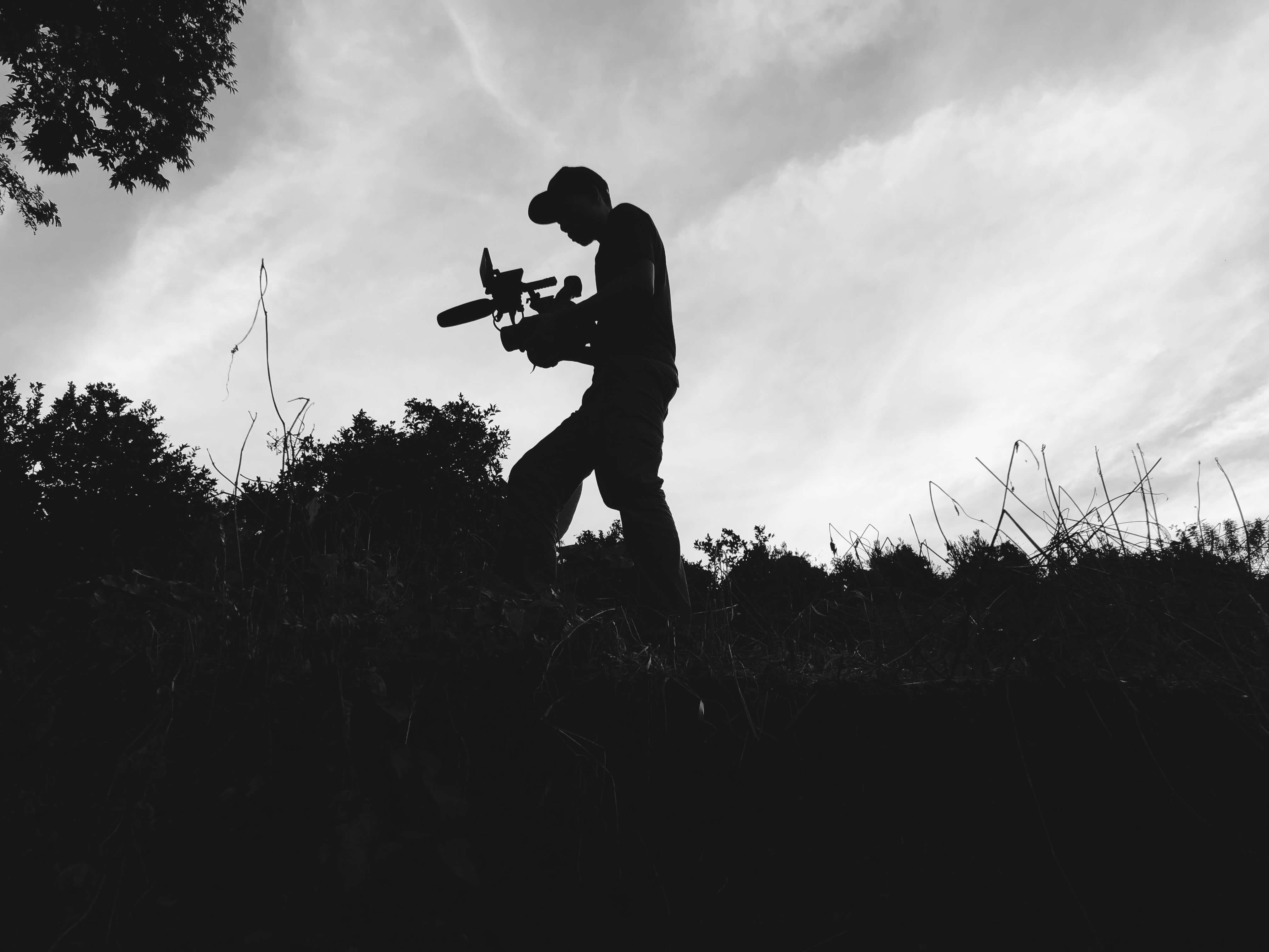 Production still from Silent Beauty. Low-angle shot of a cameraperson walking with equipment in hands, surrounded by nature. Black and white overshadowed shot.