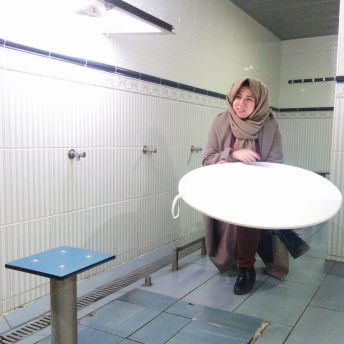 Farah Kassem sits with her legs crossed wearing a headscarf holding a large circular light reflector on her lap. In the background are tiled floors and walls and faucets. Color photograph.