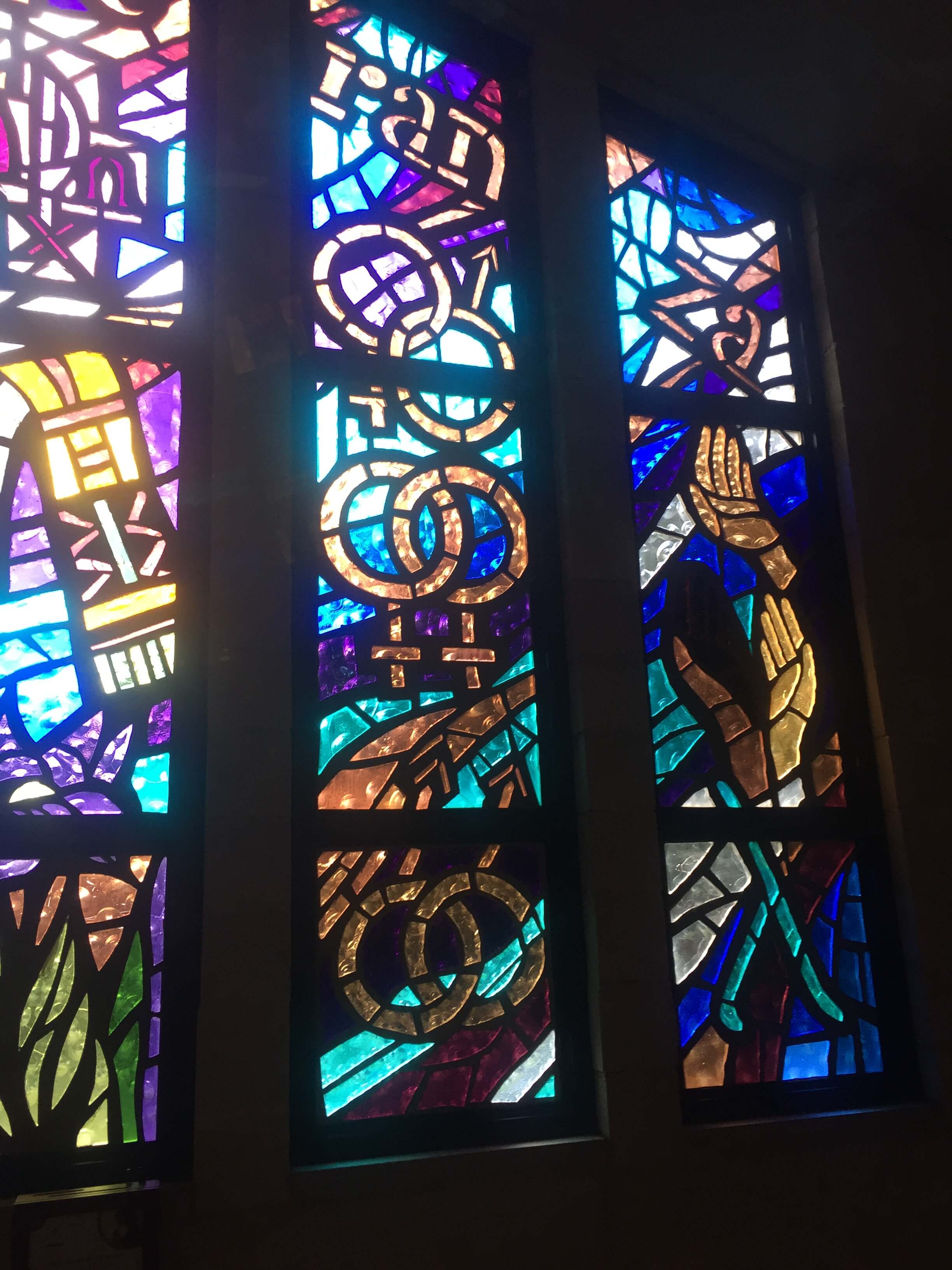 Still from Mama Bears. A stained glass window of purples, greens, and golds. There are multiple linked gender symbols, hands, and letters that read, "ran" and "za".
