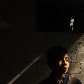 A young boy wearing a jacket in front of a bricked wall. He is in the shadow and a line of light crosses his face.