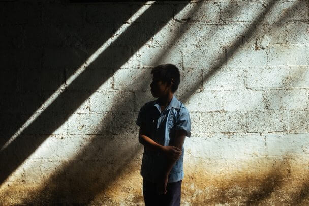 A young boy with a blue shirt looks away from the camera. He stands in front of a bricked wall. Lines of light reflect on the wall and the boy.