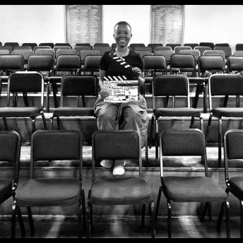 Still from Milisuthando (working title). Milisuthando is sitting amongst a few rows of chairs, smiling, and holding a film clapperboard. Black and white photo.