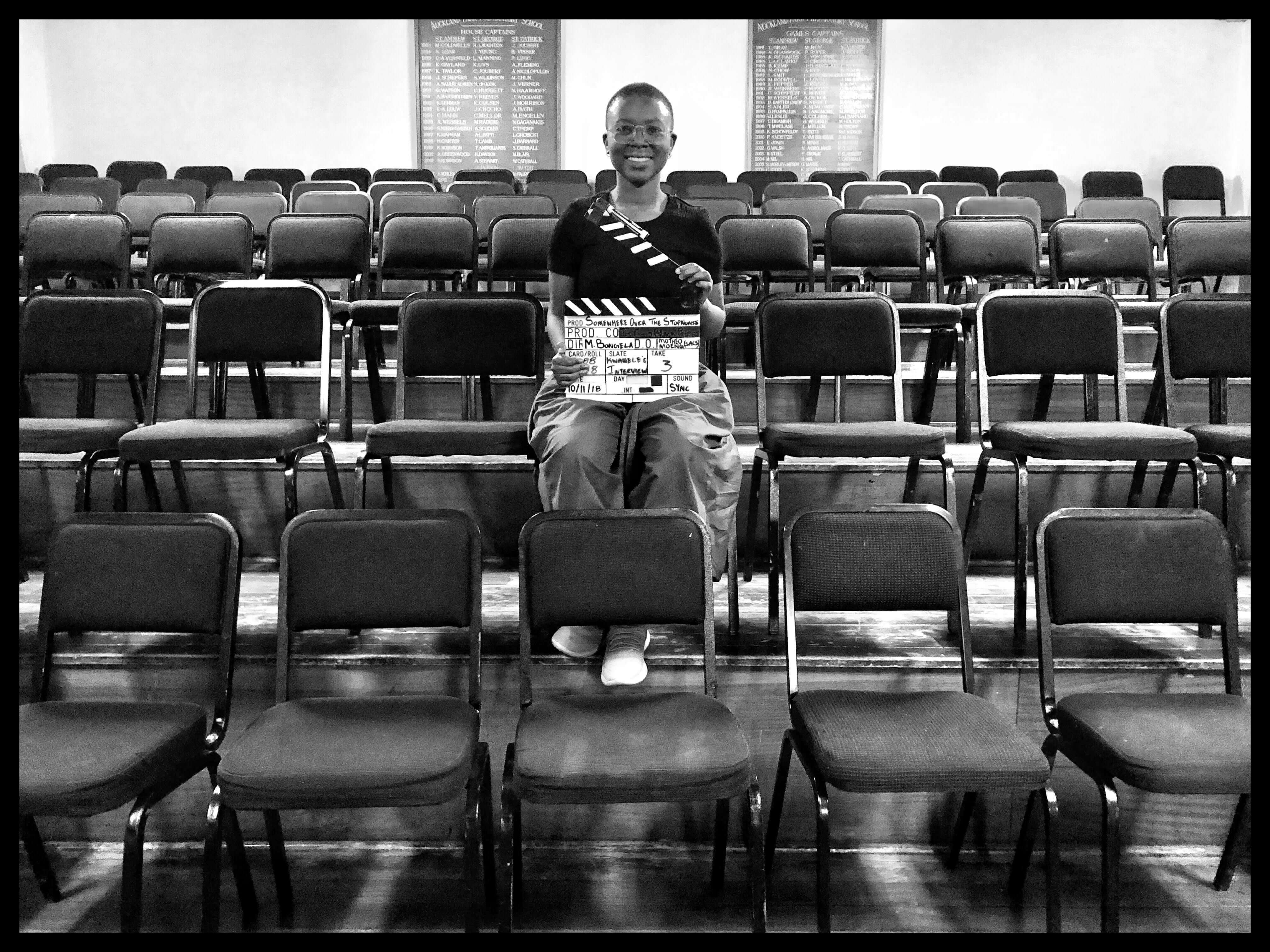 Still from Milisuthando (working title). Milisuthando is sitting amongst a few rows of chairs, smiling, and holding a film clapperboard. Black and white photo.